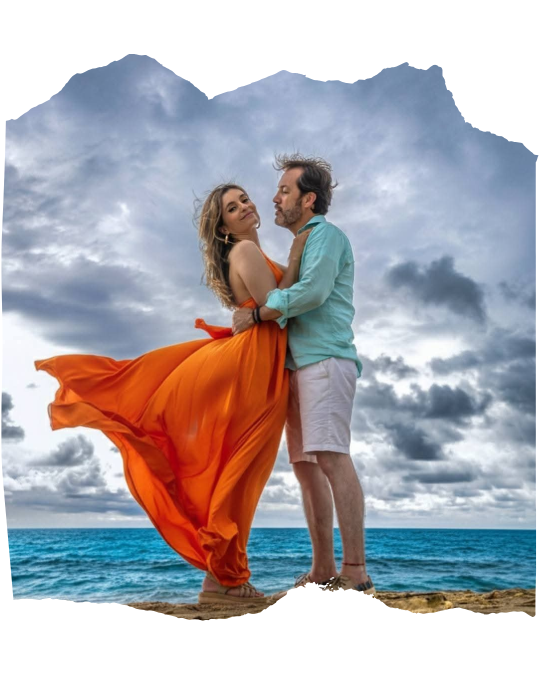Carol con vestido naranja al viento abrazada a Carlos frente al mar
