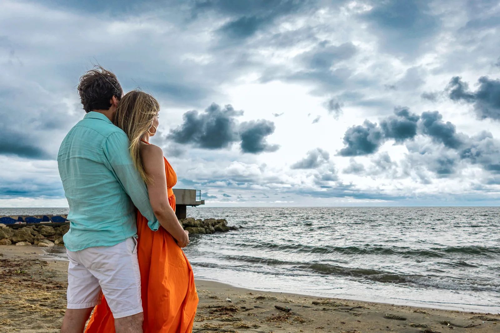 Carol y Carlos abrazados mirando el mar bajo un cielo nublado